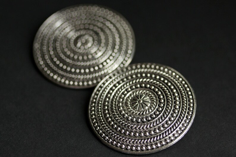 May include: Two silver circular earrings with intricate embossed designs. The earrings feature concentric circles of raised dots and a central flower-like motif.