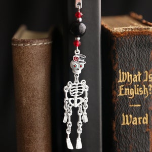May include: A silver skeleton bookmark with a red and black top hat and red eyes. The bookmark is hanging from a black bead and red crystal beads. The bookmark is hanging from a book with the title "What Is English?" by Ward.
