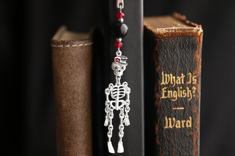 May include: A silver skeleton bookmark with a red and black top hat and red eyes. The bookmark is hanging from a black bead and red crystal beads. The bookmark is hanging from a book with the title "What Is English?" by Ward.