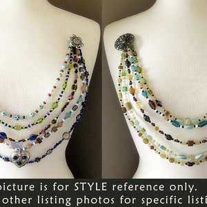 May include: Multi-strand necklace with a heart pendant. The necklace is made of various colored beads, including blue, green, red, and white. The heart pendant is silver with a blue stone in the center.