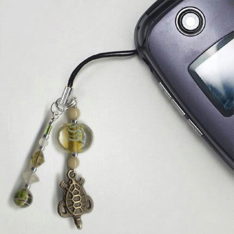 May include: A cell phone charm with a beaded chain and a turtle pendant. The beads are brown, green, and white. The turtle is made of metal and has a brown finish.