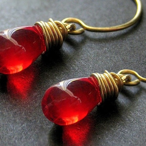以下が含まれることがあります： 赤いガラスの涙型のイヤリングのペア。各涙型の頂上に金色のワイヤーが巻かれています。イヤリングは金色のフープからぶら下がっています。