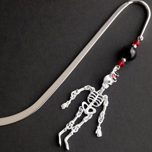 May include: Silver bookmark with a skeleton charm and black and red beads. The skeleton is white with red accents and is hanging from the bookmark.