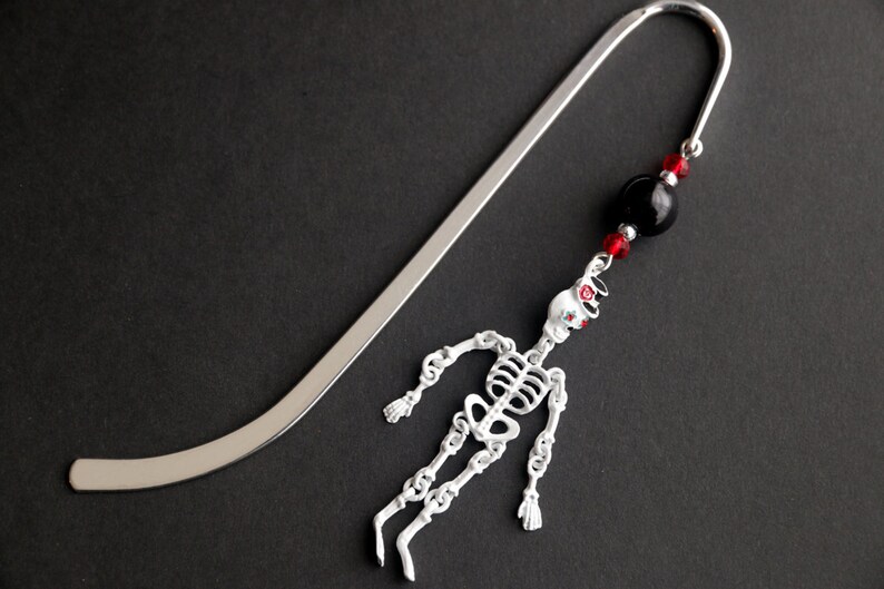 May include: Silver bookmark with a skeleton charm and black and red beads. The skeleton is white with red accents and is hanging from the bookmark.