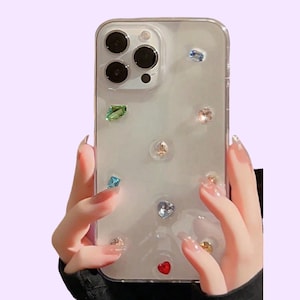 May include: A clear smartphone case featuring a variety of small, multi-colored decorative gems embedded on the back surface. The gems include various shapes and colors such as red, blue, green, and clear, arranged across the transparent cover.