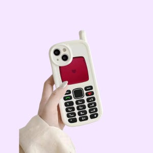 Op de afbeelding: Een witte mobiele telefoonhoes ontworpen om op een vintage mobiele telefoon met antenne te lijken. De hoes heeft een rood schermgedeelte met een klein hartpictogram en een zwart toetsenbord met witte cijfers en symbolen, inclusief een rode telefoon.
