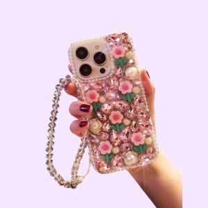 Puede incluir: Una funda para smartphone decorada con numerosos cristales rosas facetados, perlas blancas y pequeños dijes de flores rosas con tallos verdes. La funda incluye una correa de muñeca a juego hecha de cuentas transparentes y brillantes.