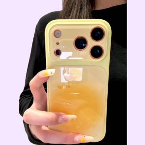 Peut inclure: Une coque de smartphone jaune pâle avec une finition brillante et une découpe rectangulaire pour l'appareil photo, dotée de trois objectifs circulaires aux bords orange. Le dos présente une section semi-transparente révélant une teinte orange.