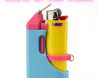 Puede incluir: Un encendedor BIC amarillo asegurado en un soporte de plástico azul claro y rosa con una mecha de cáñamo en la parte superior. El texto GLOW IN THE DARK está impreso en rosa arriba. El soporte tiene una correa rosa y un anillo de metal.