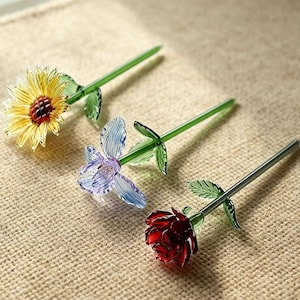 Puede incluir: Tres bolígrafos de flores de cristal: un girasol amarillo, una flor azul y morada, y una rosa roja. Cada bolígrafo tiene un tallo y hojas verdes. Los bolígrafos están colocados sobre una superficie beige texturizada.