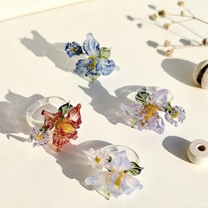 Puede incluir: Cuatro servilleteros de cristal transparente con diseños florales coloridos. Las flores son en tonos rojos, morados, azules y amarillos, con hojas verdes. Dispuestos sobre una superficie blanca.