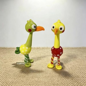 May include: Two whimsical glass bird figurines. One is green with yellow polka dots and a striped leg. The other is yellow with a red polka dot body and striped legs. Both have orange beaks and large eyes.