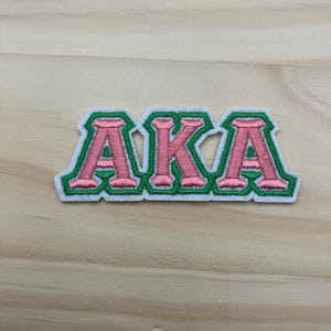 May include: Embroidered patch featuring the letters "AKA" in pink with a green border and white backing. The patch is displayed on a light wood grain surface. The letters are in a bold, stylized font.