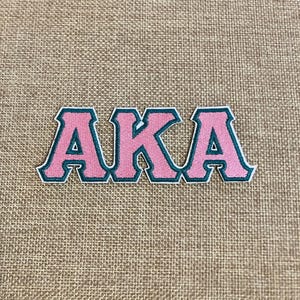 May include: Pink and green embroidered patch with the letters "AKA" in a bold, collegiate font. The letters are pink with a green outline and a white border. The patch is likely for a sorority or organization.