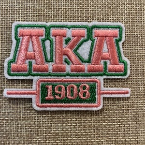 May include: Embroidered patch with the letters "AKA" in pink with green and white detailing. Beneath the letters is a green rectangle with the number "1908" in pink. The patch is set against a tan fabric background.