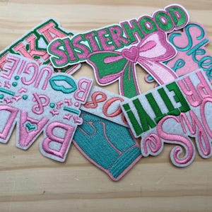May include: A collection of embroidered patches in various shapes and sizes. The patches feature phrases like "SISTERHOOD," "PRETTY," and "BAD," in pink, green, and teal thread. The patches are on a light brown wooden surface.
