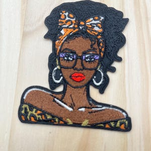 May include: Embroidered patch featuring a Black woman with dark curly hair, wearing glasses, hoop earrings, and a bow. The bow and headband have an orange and black leopard print. Her lips are red, and her skin is brown. The patch has a black border.