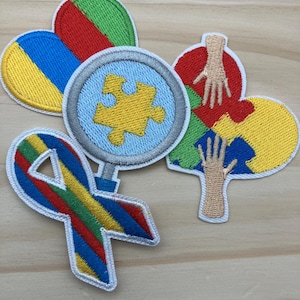 May include: Embroidered patches in various shapes and colors, including hearts, a ribbon, and a magnifying glass with a puzzle piece. The patches feature a rainbow of colors and are likely related to autism awareness.