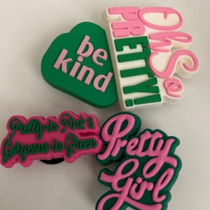 May include: Four colourful shoe charms with text. One green heart-shaped charm says "be kind" in pink. Other charms say "Oh So Pretty!", "Pretty in Pink & Gorgeous in Green", and "Pretty Girl" in pink and green.