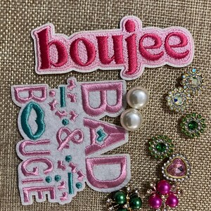 以下が含まれることがあります： 刺繍パッチと装飾ジュエリー。パッチには「boujee」と「boujee & bad」の文字がピンクとティールで表示されています。ジュエリーには、パールのイヤリング、ピンク、グリーン、クリアのラインストーンと宝石がちりばめられたブローチが含まれています。
