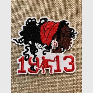 May include: Embroidered patch featuring a profile of a Black woman with black hair and a red head wrap. The patch includes the numbers "1913" in red, with a red figure between the numbers. The patch is outlined in white.
