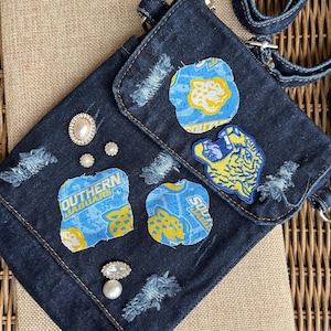 May include: A dark denim crossbody bag with a flap closure, decorated with patches displaying a blue and yellow jaguar logo and the text "SOUTHERN JAGUARS". The bag features pearl and rhinestone embellishments and a denim strap.