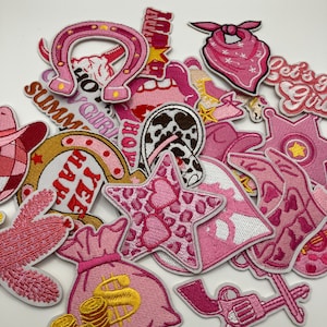 May include: A collection of pink embroidered patches with Western and summer themes. Patches include a horseshoe, a star, a money bag, a bandana, a sheriff's badge, and a revolver. Some patches have text like "Summer Girl" and "Yee Haw".