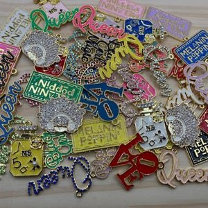 Könnte beinhalten: Eine Sammlung farbenfroher, goldfarbener Charms mit verschiedenen Designs und Texten. Die Charms enthalten die Wörter "Queen", "Melanin Poppin" und "N5". Einige Charms weisen ein Parfümflaschendesign auf, andere haben ein funkelndes, juwelenbesetztes Aussehen.