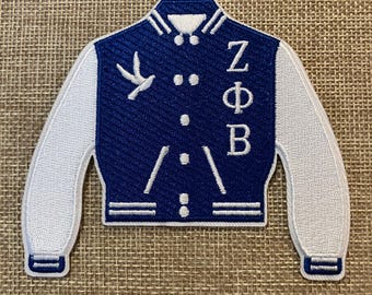 Peut inclure: Écusson brodé bleu et blanc en forme de veste universitaire. La veste présente des manches blanches, un emblème d'oiseau et les lettres grecques Z, Φ et B. L'écusson est sur un fond de tissu beige.