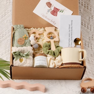 May include: A baby gift box with various items. Inside, there's a welcome baby card, a green drawstring bag, a wooden rattle, a candle, a mug, and a pair of socks. The box is brown, and the items are in shades of green, white, and beige. The text on the card reads 'Welcome baby' and 'Feel better soon, my friend In Your Consumers buddy Wendy'. There is also a wooden toy car.