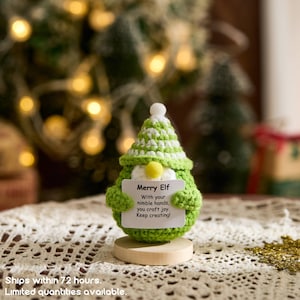 May include: A handmade green and white crocheted elf figurine with a yellow nose and a white paper sign that reads "Merry Elf". The sign has text that says "With your nimble hands, you craft joy. Keep creating!"
