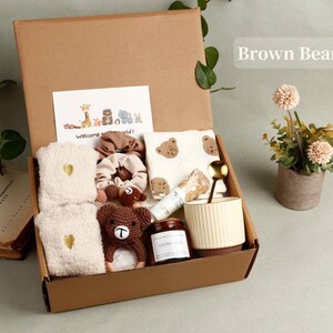 Pode incluir: Uma caixa de presente de cart&atilde;o castanho cheia de artigos para beb&eacute;, incluindo um urso de croch&eacute;, toalhas, uma caneca e um cart&atilde;o que diz "Welcome world!". Uma pequena planta em vaso est&aacute; ao lado da caixa.