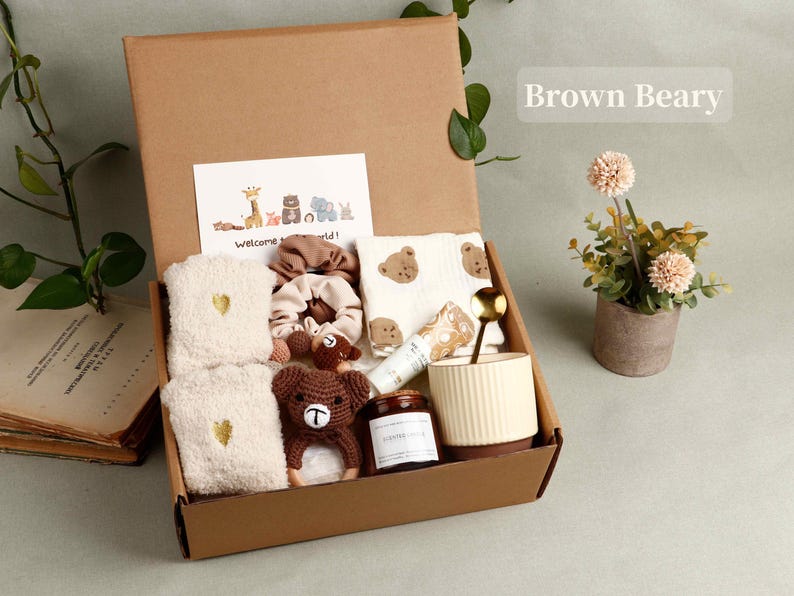 Pode incluir: Uma caixa de presente de cart&atilde;o castanho cheia de artigos para beb&eacute;, incluindo um urso de croch&eacute;, toalhas, uma caneca e um cart&atilde;o que diz "Welcome world!". Uma pequena planta em vaso est&aacute; ao lado da caixa.