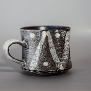 May include: A black and white ceramic mug with a geometric pattern. The mug has a handle and a rounded bottom.