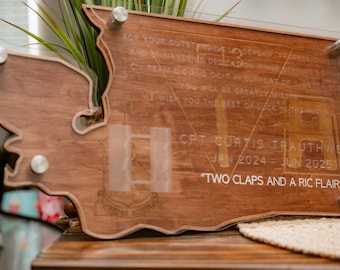 May include: A wooden plaque with a state outline, engraved with the text "FOR YOUR OUTSTANDING LEADERSHIP PROFESSIONALISM AND UNWAVERING DEDICATION WHILE SERVING C-TEAM DIG AND OC/T IN SUPPORT OF 95 YOU WILL BE GREATLY MISSED WE WISH YOU THE BEST OF LUCK IN THE FUTURE CPT. CURTIS TRAUTHWEIN JAN 2024-JUN 2025 "TWO CLAPS AND A RIC FLAIR""