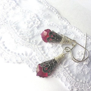 Puede incluir: Un par de pendientes colgantes con gemas facetadas de color rojo intenso. Los pendientes presentan capuchones de metal oscuro adornados con detalles de filigrana y están suspendidos de ganchos plateados. Los pendientes descansan sobre encaje blanco.