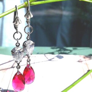 May include: Silver-tone drop earrings featuring clear crystal elements and vivid, teardrop-shaped, fuchsia gems. The earrings are suspended from a floral-shaped top. The earrings are displayed against a bright, natural background.