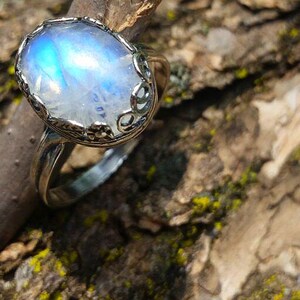 May include: A silver ring with a large, oval, iridescent moonstone. The stone has a blue and white glow. The ring is set on a piece of textured wood.