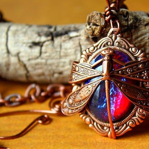 K&ouml;nnte beinhalten: Eine kupferfarbene Libellen-Anh&auml;nger-Halskette. Der Anh&auml;nger zeigt ein Libellen-Design mit detaillierten Fl&uuml;geln und einem zentralen Stein mit roten und blauen Farbt&ouml;nen. Die Halskette ist ebenfalls kupferfarben und liegt auf einem Holzscheit.