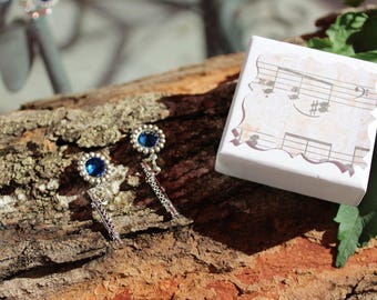 May include: A pair of silver earrings with blue gemstone centers and dangling accents, displayed on a piece of wood. A white box with a musical notes design sits nearby. The earrings and box are set against a natural background.