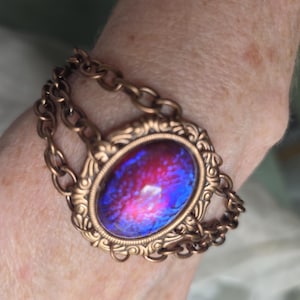 Puede incluir: Una pulsera de tono bronce con un cabujón ovalado iridiscente en tonos rojos y azules. El cabujón está engastado en un marco decorativo, y la pulsera tiene un diseño de eslabones de cadena.