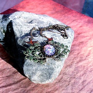 Puede incluir: Un collar artesanal con un colgante central redondo e iridiscente en tonos morados y azules. El colgante est&aacute; enmarcado por una ornamentaci&oacute;n met&aacute;lica color bronce con detalles de hojas y vides. Dos peque&ntilde;os amuletos de p&aacute;jaros rojos flanquean el colgante. El collar descansa sobre una piedra gris.