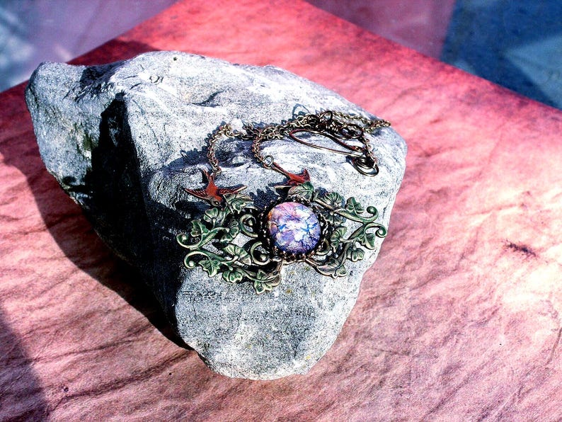 Puede incluir: Un collar artesanal con un colgante central redondo e iridiscente en tonos morados y azules. El colgante est&aacute; enmarcado por una ornamentaci&oacute;n met&aacute;lica color bronce con detalles de hojas y vides. Dos peque&ntilde;os amuletos de p&aacute;jaros rojos flanquean el colgante. El collar descansa sobre una piedra gris.
