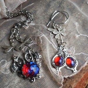 Könnte beinhalten: Eine silberfarbene Halskette und ein passendes Ohrring-Set. Die Halskette hat einen Anhänger mit einem rot-blauen Stein, und die Ohrringe haben ähnliche Steine. Der Schmuck liegt auf einem Spitzengrund.