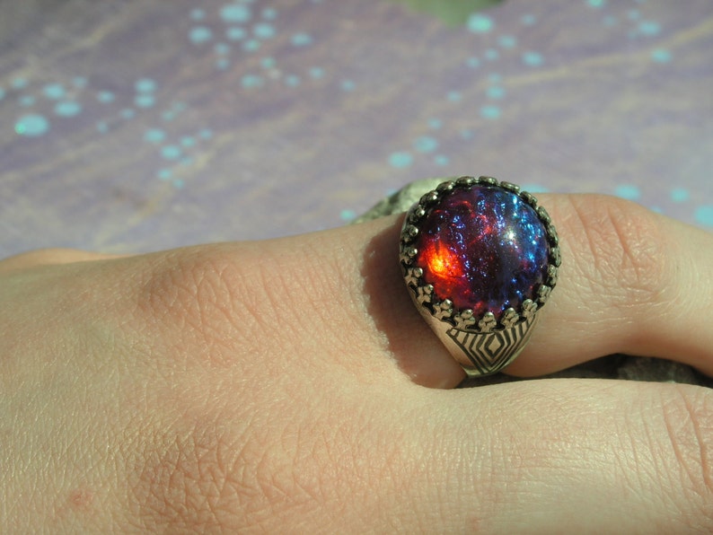May include: A silver ring with a large, round, red and blue iridescent gemstone set in a detailed silver bezel.