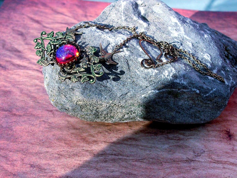 Puede incluir: Un collar de tono bronce con un colgante grande, redondo e iridiscente en tonos morados y rojos. El colgante est&aacute; engastado en un marco decorativo con hojas y p&aacute;jaros. La cadena descansa sobre una piedra gris sobre un fondo rosa.