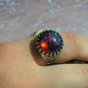 May include: A silver ring with a large, round, red and blue iridescent gemstone set in a decorative silver bezel.