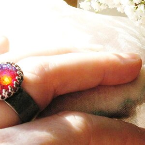 May include: A silver ring with a red and purple gemstone set in a decorative silver bezel. The ring is worn on a finger.