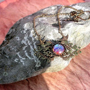 Puede incluir: Un collar de tono bronce con una piedra redonda iridiscente central. El collar presenta elementos decorativos de p&aacute;jaros y dise&ntilde;os florales. La cadena y el colgante descansan sobre una roca gris sobre un fondo rosa.