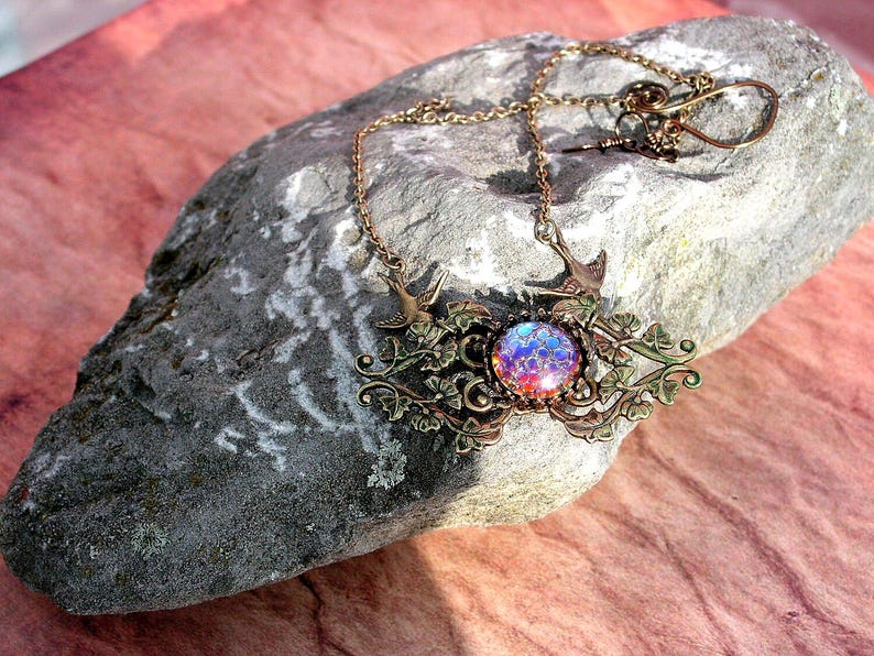 Puede incluir: Un collar de tono bronce con una piedra redonda iridiscente central. El collar presenta elementos decorativos de p&aacute;jaros y dise&ntilde;os florales. La cadena y el colgante descansan sobre una roca gris sobre un fondo rosa.
